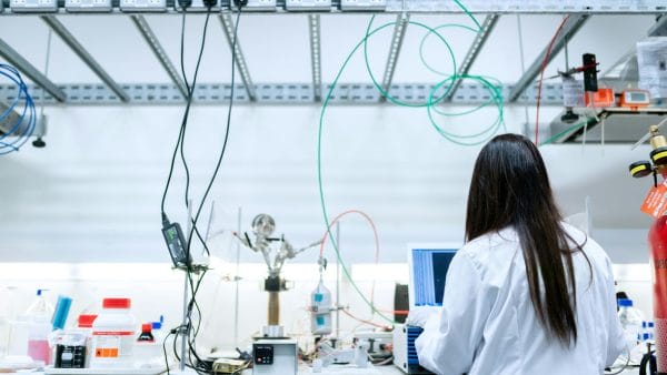 A female scientist with long dark hair, wearing a white lab coat, works at a cluttered optics and photonics research bench. The setup features numerous green, red, and black optical fibres looped overhead, precision mounts, a laser source, power supplies, and a computer monitor displaying data, in a bright, modern laboratory environment.