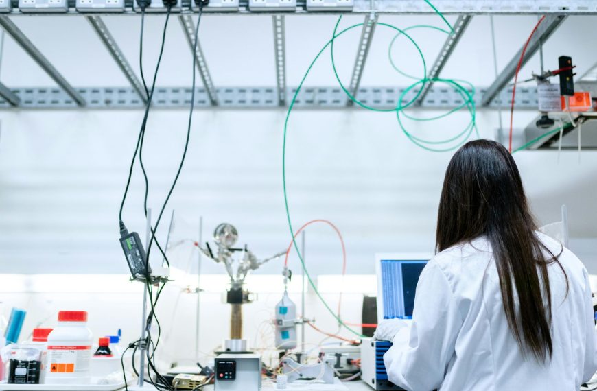 A female scientist with long dark hair, wearing a white lab coat, works at a cluttered optics and photonics research bench. The setup features numerous green, red, and black optical fibres looped overhead, precision mounts, a laser source, power supplies, and a computer monitor displaying data, in a bright, modern laboratory environment.