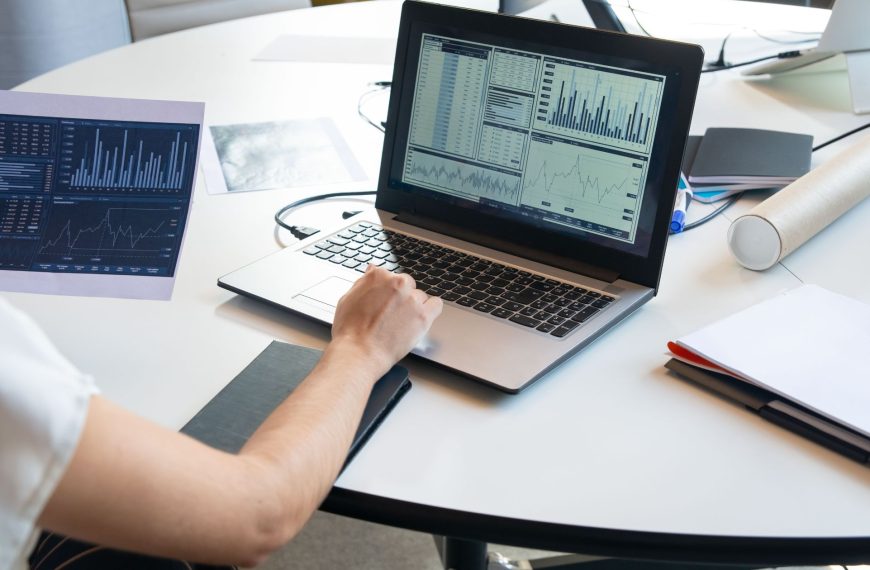 A business professional reviewing financial data and performance metrics on a laptop dashboard and printed charts.