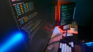A close-up of a computer monitor displaying lines of software code on a dual-screen desktop setup with atmospheric lighting, representing application development.