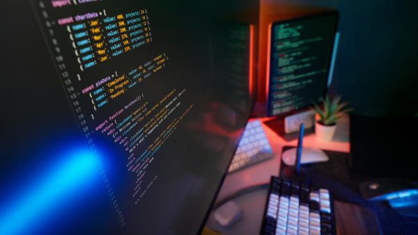 A close-up of a computer monitor displaying lines of software code on a dual-screen desktop setup with atmospheric lighting, representing application development.