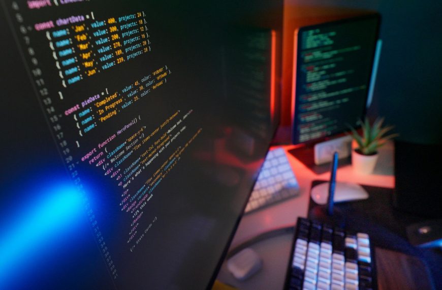 A close-up of a computer monitor displaying lines of software code on a dual-screen desktop setup with atmospheric lighting, representing application development.