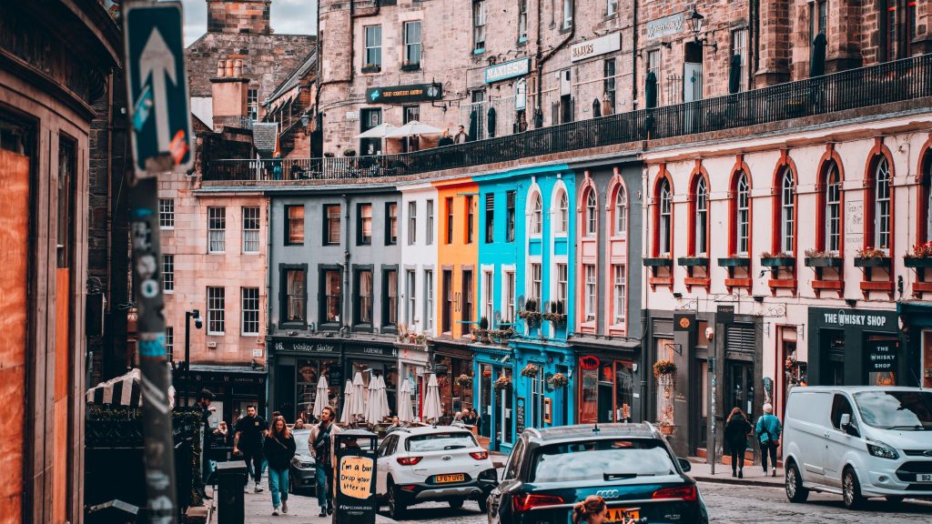 A bustling, curved cobblestone street in Edinburgh lined with vibrant, colourful storefronts, pedestrians, and local traffic.