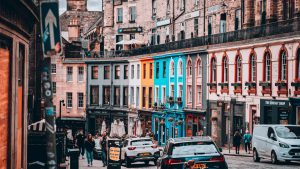 A bustling, curved cobblestone street in Edinburgh lined with vibrant, colourful storefronts, pedestrians, and local traffic.