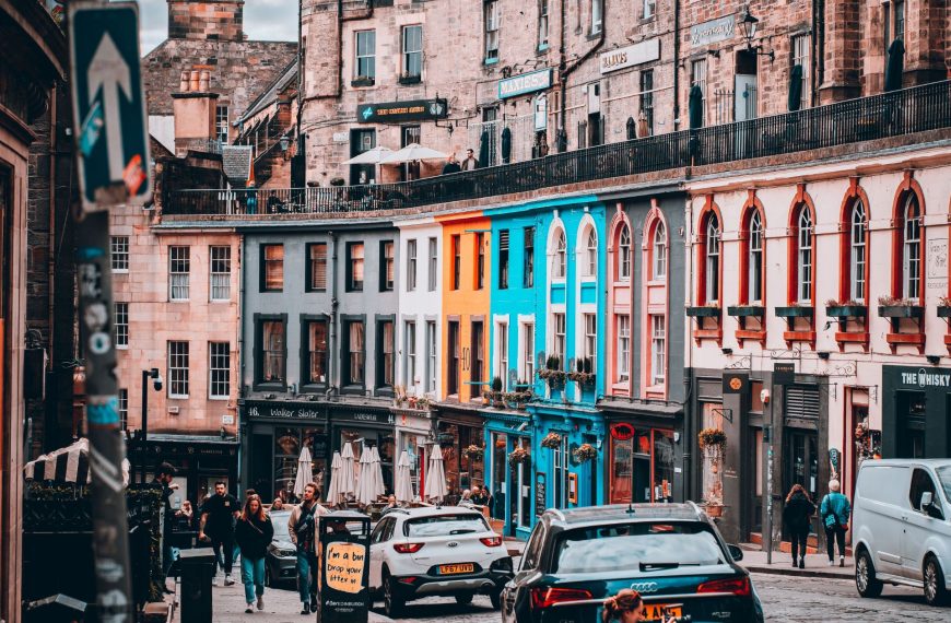 A bustling, curved cobblestone street in Edinburgh lined with vibrant, colourful storefronts, pedestrians, and local traffic.