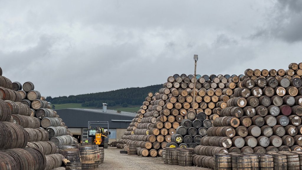 Large-scale outdoor inventory storage showing hundreds of wooden barrels stacked high outside an industrial facility, with a forklift handling materials in the background.