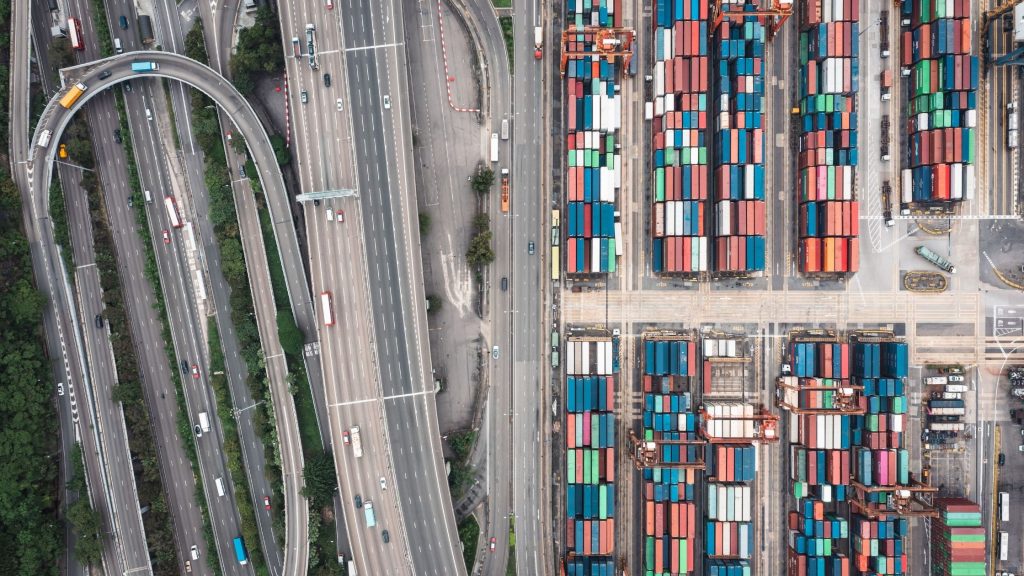 An aerial photograph showing a complex multi-lane highway interchange situated immediately adjacent to a dense, highly organised shipping container terminal.