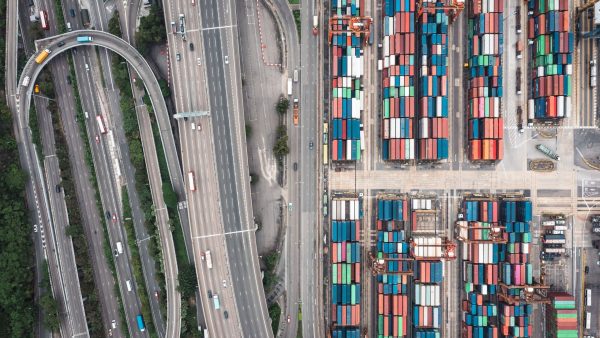 An aerial photograph showing a complex multi-lane highway interchange situated immediately adjacent to a dense, highly organised shipping container terminal.