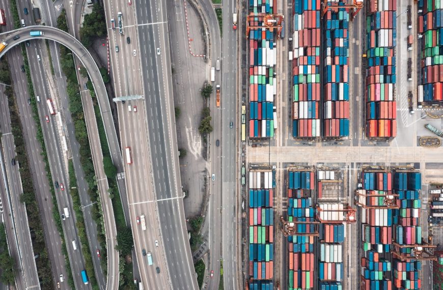 An aerial photograph showing a complex multi-lane highway interchange situated immediately adjacent to a dense, highly organised shipping container terminal.