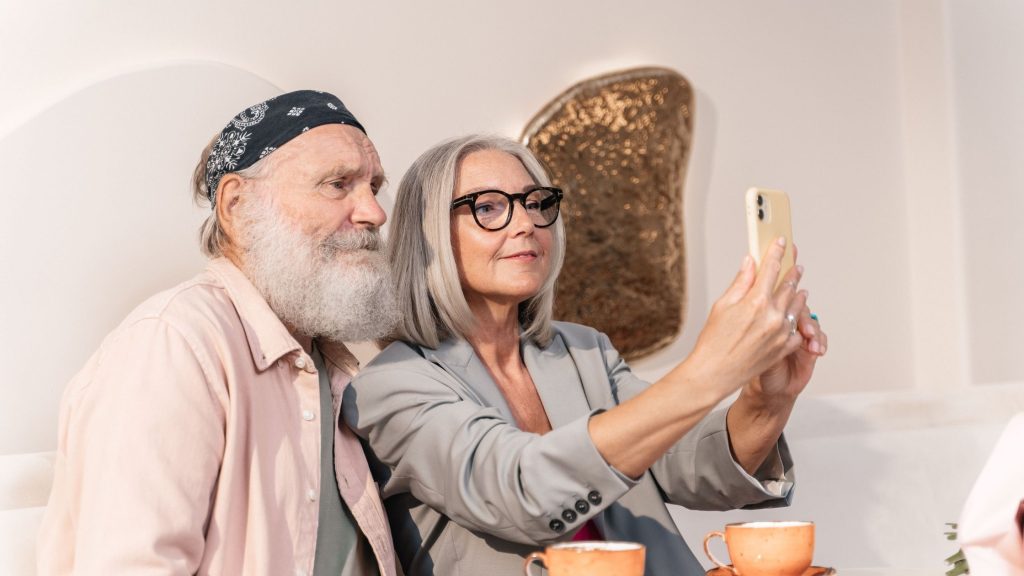A stylish older couple taking a selfie with a smartphone while sitting at a cafe table, illustrating authentic age-based marketing and everyday digital engagement.
