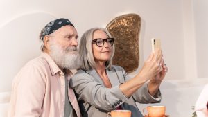 A stylish older couple taking a selfie with a smartphone while sitting at a cafe table, illustrating authentic age-based marketing and everyday digital engagement.