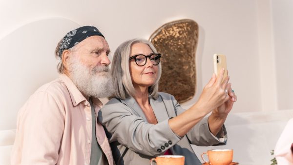 A stylish older couple taking a selfie with a smartphone while sitting at a cafe table, illustrating authentic age-based marketing and everyday digital engagement.