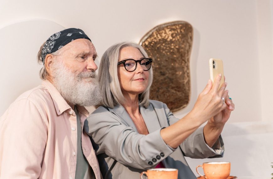 A stylish older couple taking a selfie with a smartphone while sitting at a cafe table, illustrating authentic age-based marketing and everyday digital engagement.